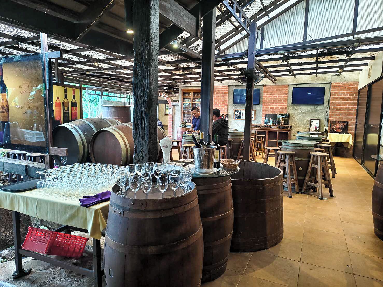 A rustic interior with large wooden barrels, a table with wine glasses, and a brick wall.