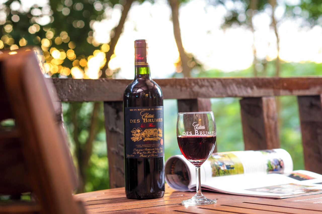 A bottle of wine and a glass on a wooden table with a magazine, set outdoors with a blurred natural background.