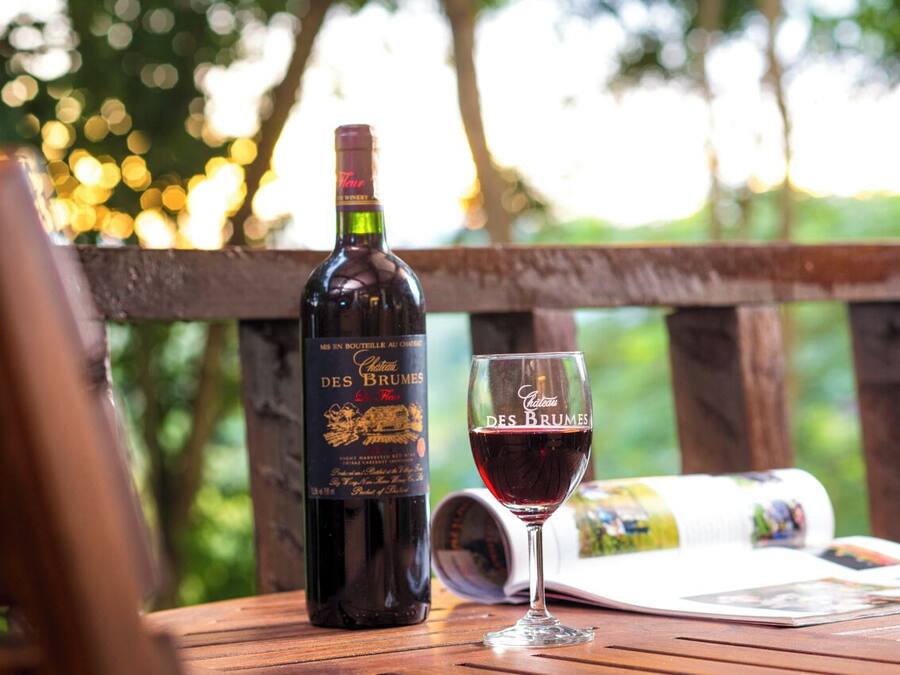 A bottle of wine and a glass on a wooden table with a magazine, set outdoors with a blurred natural background.