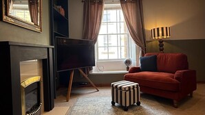 A living room with a fireplace, a television, a red sofa, a striped ottoman, and a lamp. - Charming Grade 2 Listed Apartment in Alnwick town centre with castle views. (Alnwick)