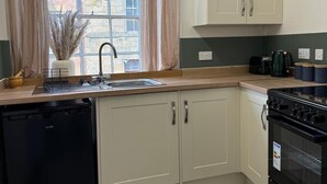 A compact kitchen with a dishwasher, stove, and a window with curtains. - Charming Grade 2 Listed Apartment in Alnwick town centre with castle views. (Alnwick)