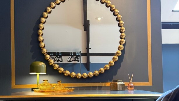 A room with a dark blue wall, a round mirror framed with gold, a small table with a lamp, and a chair. - Charming Grade 2 Listed Apartment in Alnwick town centre with castle views. (Alnwick)