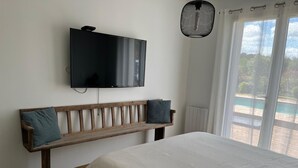 A bedroom with a bed, a wooden bench with cushions, a TV mounted on the wall, and a window with curtains. - House with swimming pool near Contis beach, cycle path and all amenities (Saint-Julien-en-Born)