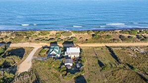 Vista aérea de una propiedad costera con una carretera que lleva a una casa de techo verde, ubicada en una playa de arena con vegetación y el océano al fondo.
