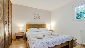 Ein Schlafzimmer mit einem Holz-Kopfteil, einer blumigen Tagesdecke, zwei Nachttischlampen und einem Fenster mit Blick auf Grünflächen.