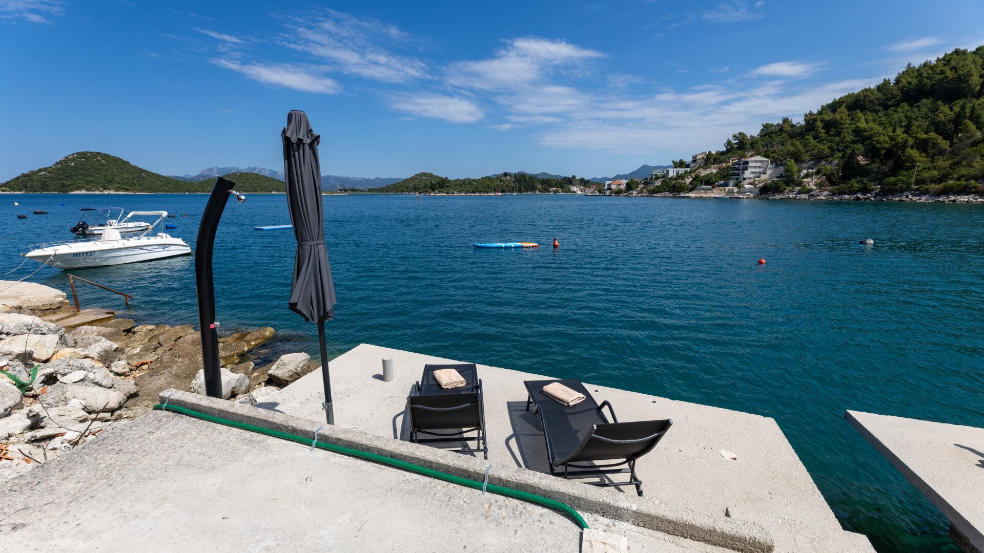 Um terraço à beira-mar com duas cadeiras de descanso, um guarda-sol e um barco atracado nas proximidades.