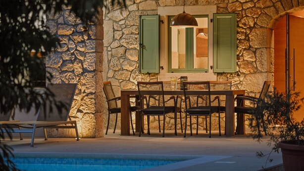 Un muro de piedra con una piscina, una mesa con sillas y una lámpara colgante.