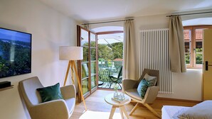 Ein modernes Wohnzimmer mit großem Fernseher, zwei Sesseln, einer Couch, einem kleinen Tisch mit Gläsern und Blick auf einen Garten durch eine Glastür.