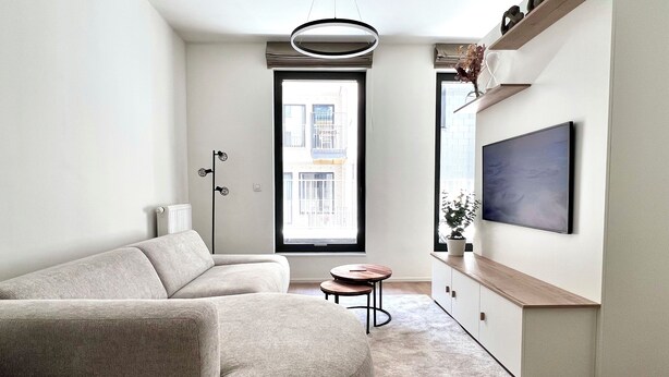 Un salon moderne avec un canapé, une petite table ronde, un lampadaire, un t éléviseur et une grande fenêtre donnant sur des immeubles.