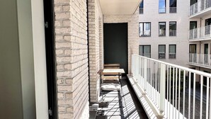 Un balcon étroit avec une balustrade blanche, un mur de briques d’un côté et une vue sur un immeuble à plusieurs étages doté de balcons.