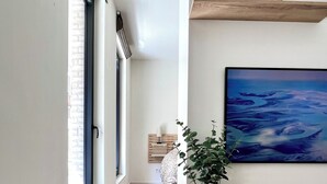 Un couloir avec un plancher en bois, un mur blanc, une fenêtre, un lit, une plante, une étagère avec des objets décoratifs et un tableau.