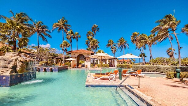 A clear blue pool with lounge chairs and umbrellas, surrounded by palm trees and a building with a red roof.