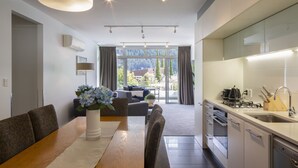 Eine moderne Küche mit Einbauherd, Spüle und Geschirrspüler. Ein Essbereich mit Holztisch und Stühlen sowie eine Couch laden zum Verweilen ein. Große Fenster mit Vorhängen bieten Blick auf viel Grün.