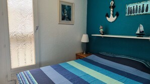 Ein Schlafzimmer mit gestreifter Bettdecke, einem gerahmten Bild an der Wand, einem kleinen Regal mit Dekoration und einem Fenster mit einem gemusterten Rollo.