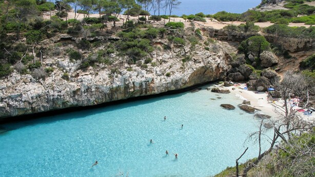 Una playa costera con agua turquesa cristalina, terreno rocoso y vegetación escasa.