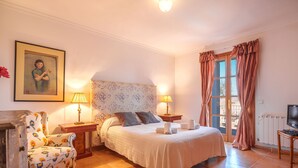 Un dormitorio con cama, dos mesitas de noche con lámparas, un sillón estampado con motivos florales, un cuadro en la pared y una ventana con cortinas.