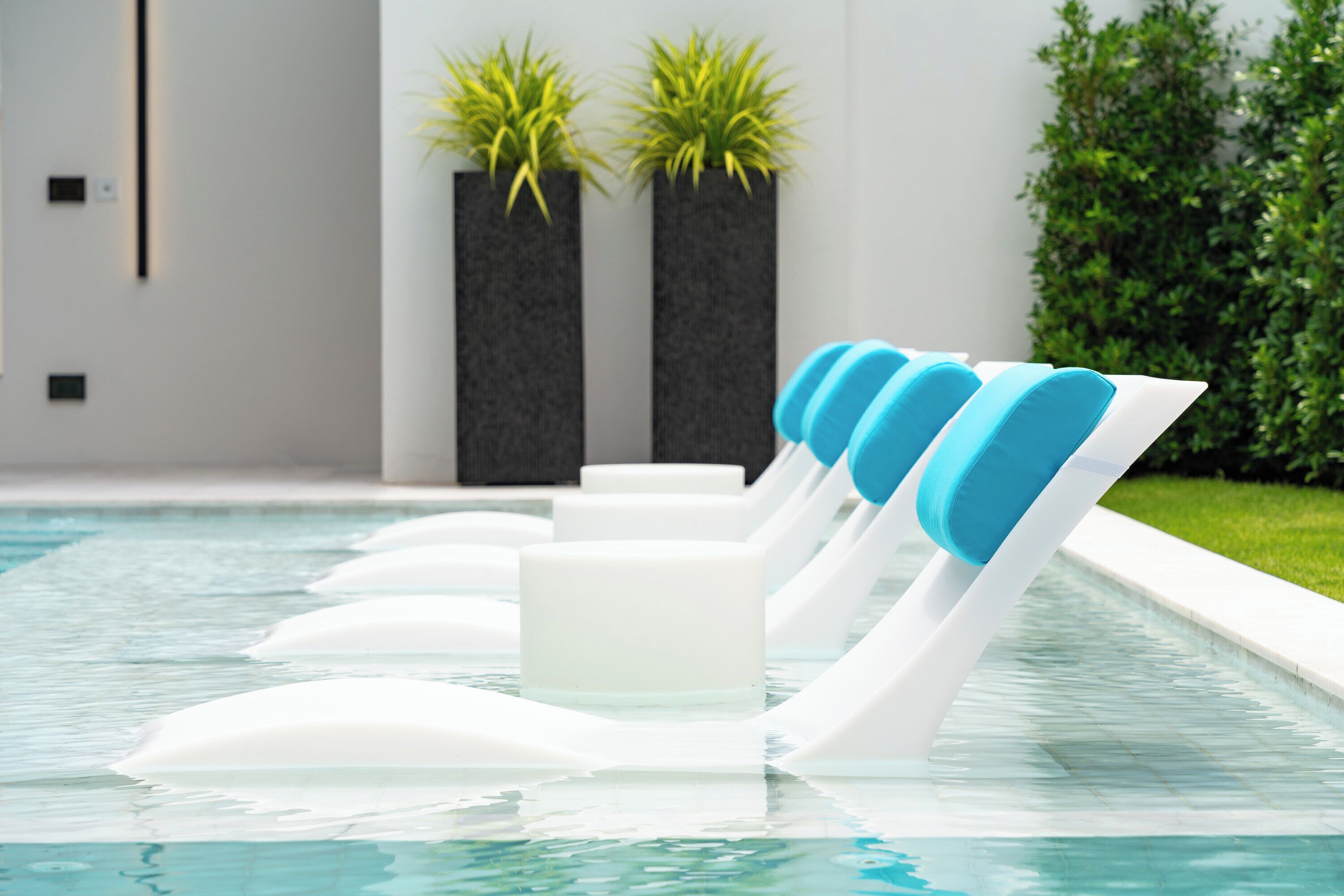 Una piscina interior moderna con tumbonas blancas y azules flotando, macetas negras con plantas verdes y un césped bien cuidado al fondo.