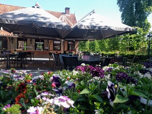 Sit and enjoy a meal under the shade of large umbrellas, surrounded by vibrant flowering plants and a charming brick building.
