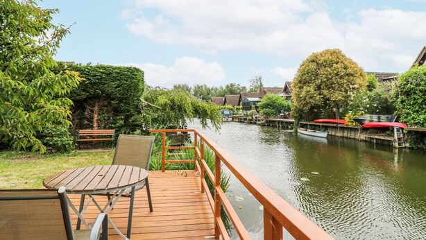 Une terrasse en bois avec une table et des chaises, donnant sur une rivière paisible, avec des bateaux et des maisons en arrière-plan.