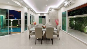 A modern dining area with a glass table and chairs, a pool visible through the glass, and an air conditioning unit on the wall. - Luxury Pool Villa A14 3BR/8 Persons (Muang Pattaya)