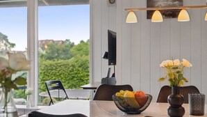 Un coin repas comprenant une table, des chaises, un saladier de fruits, un vase de fleurs et un luminaire suspendu.