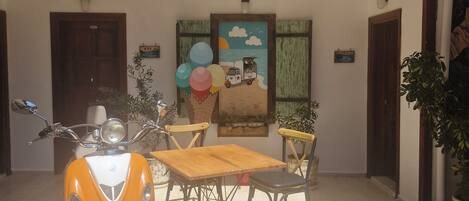 A covered outdoor area with a scooter, a table, chairs, and a painting on the wall.