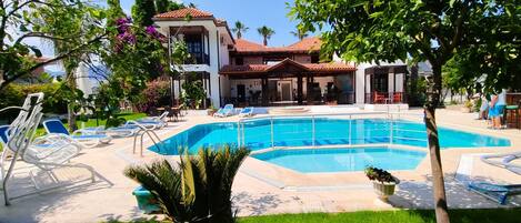 A swimming pool with lounge chairs, a covered seating area, and a building with a tiled roof.