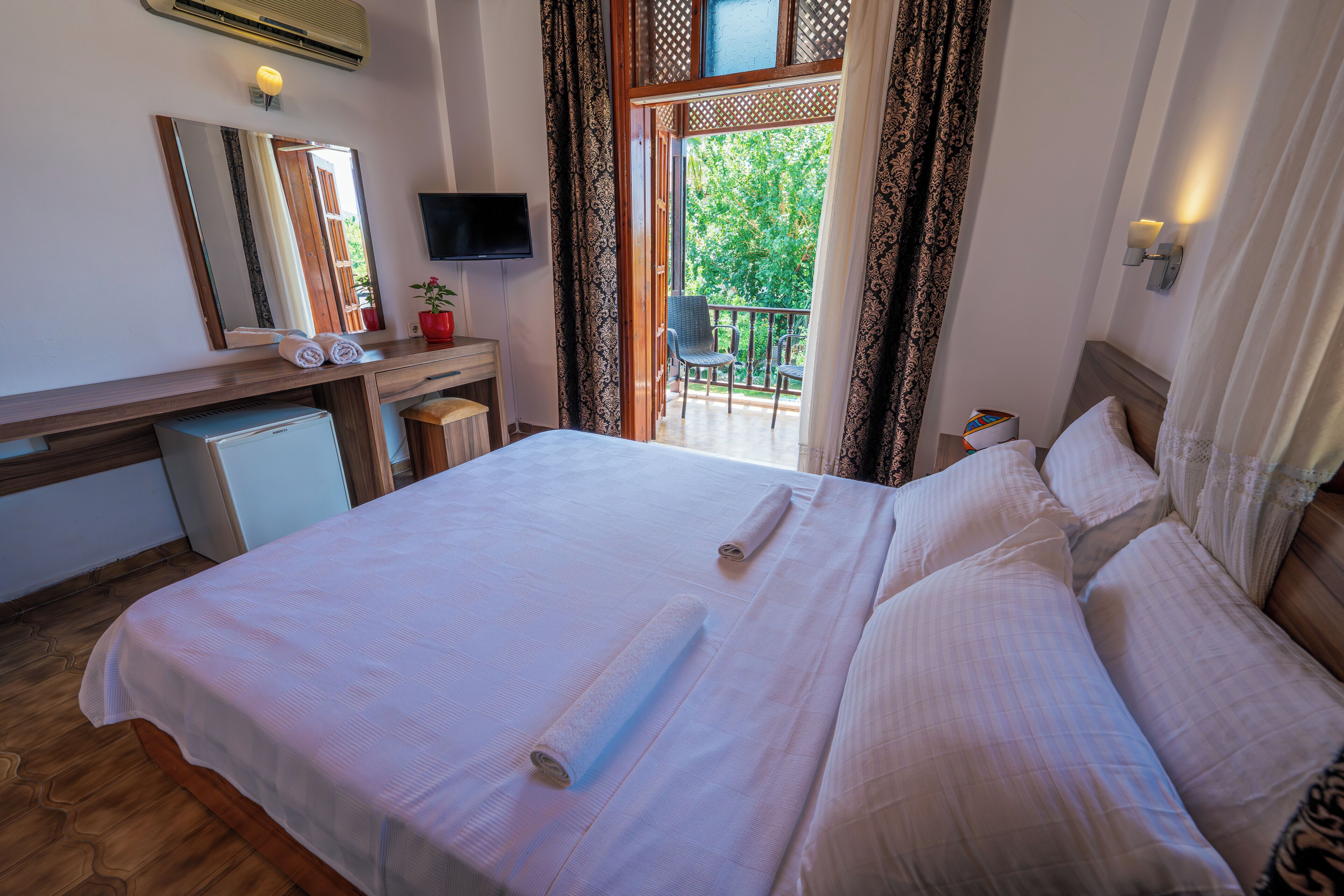 A hotel room with a large bed, a desk with a computer, a TV, and a balcony with a chair.