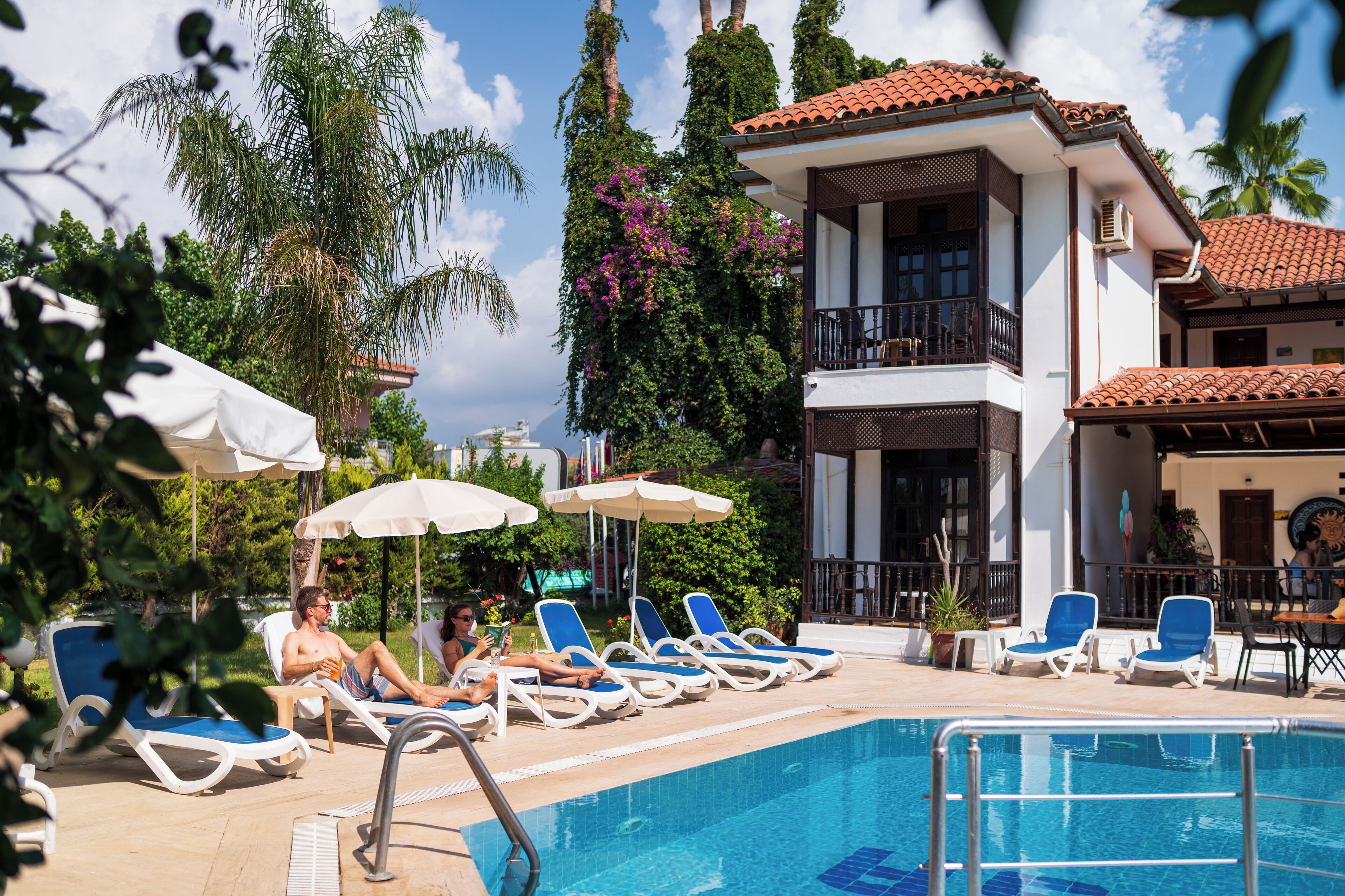 Zona de piscina con sillones de descanso y sombrillas, rodeada de vegetación y un edificio con balcón.