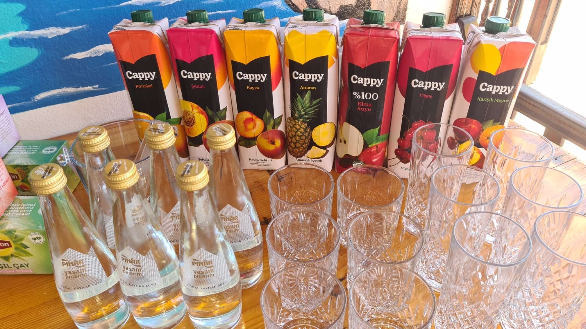Diversas embalagens de suco Cappy e garrafas da marca Pinar sobre uma mesa de madeira, com copos vazios à frente.