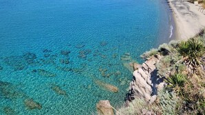 A coastal view with clear turquoise waters, rocky shoreline, and buildings on the right.
