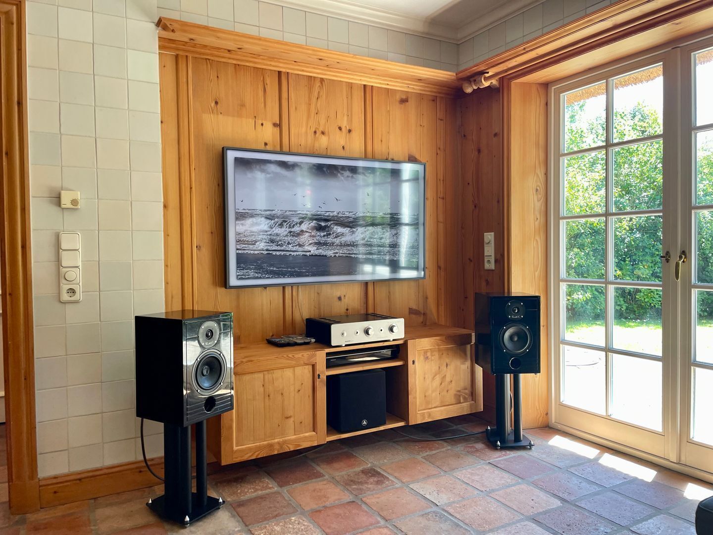 A room with a wooden paneled wall, a mounted flat-screen TV, a stereo system, and a large window with a view of greenery.