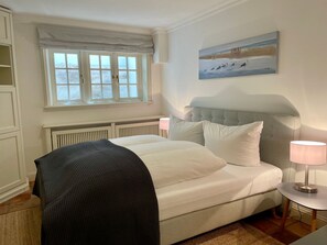 A bedroom with a bed, bedside lamps, a window with a view, and a painting on the wall. - Landhaus Sylt Glück - Holiday home Sylt Glück Morsum, 135sqm for up to 5 people (Sylt-Ost)