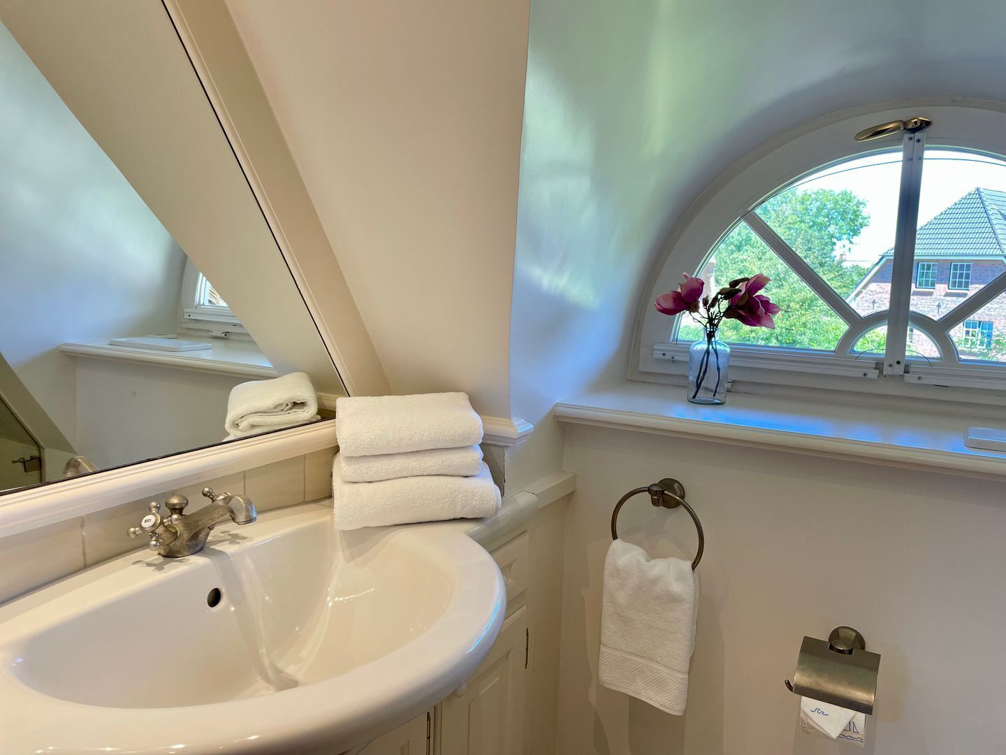 A bathroom with a white sink, a mirror, a window with a view of a house, and a vase with flowers.