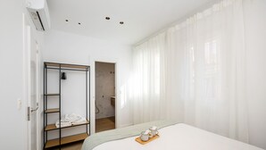 A hotel room with a bed, a wooden shelf with towels, a bathroom visible through an open door, and sheer curtains on the windows. - ChezmoiHomes Realejo Boutique Terrace (Granada)