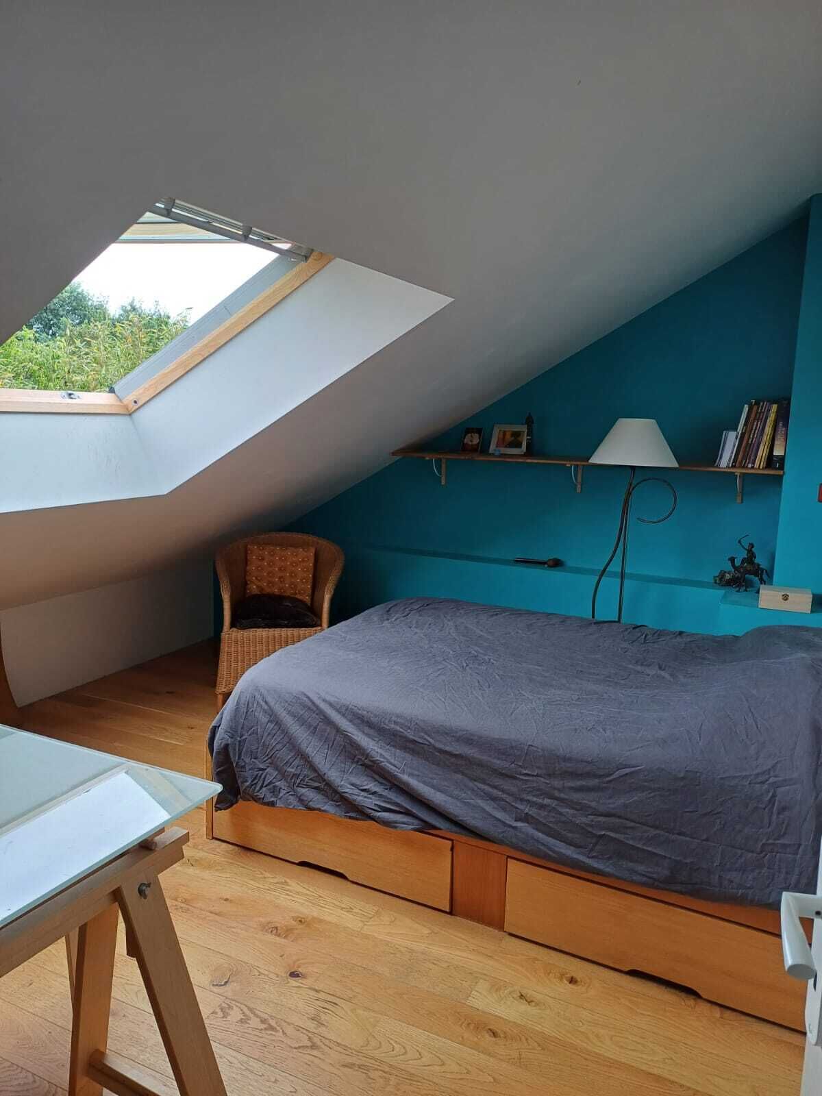 Un dormitorio con techo inclinado, un tragaluz, una cama con colcha gris, una silla, una mesita y una estantería con libros y una lámpara.