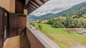 Un balcón con piso a cuadros, puertas de madera y vistas a un paisaje verde con montañas y un pueblo.