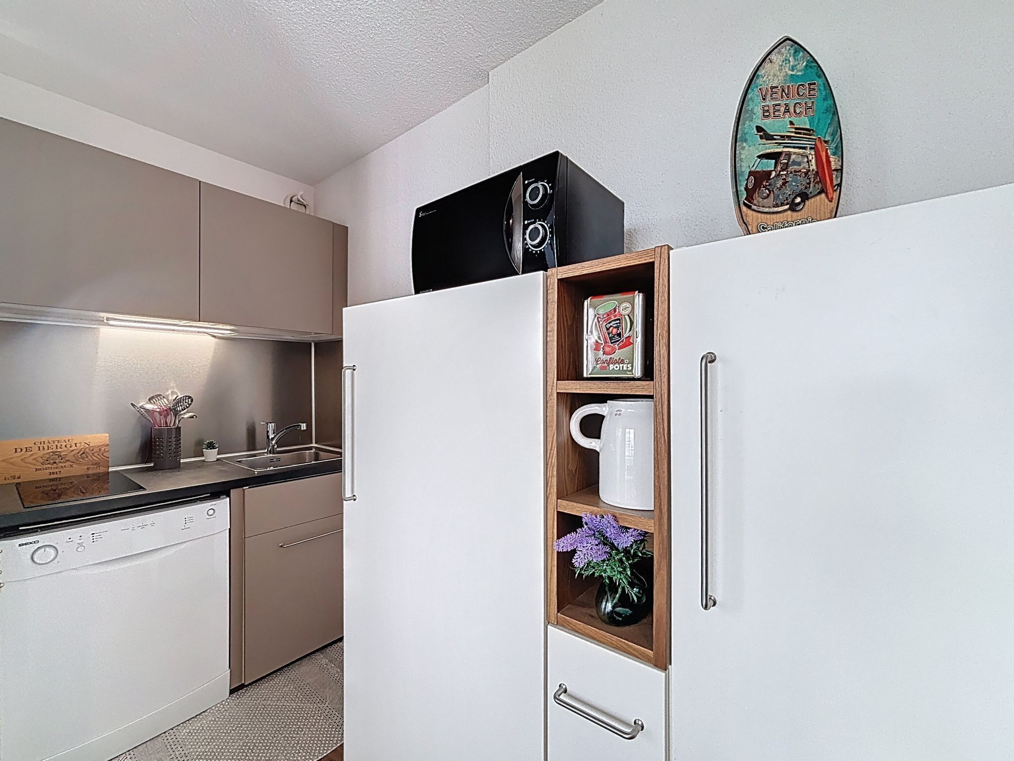 A kitchen with a built-in dishwasher, a wooden shelf with a mug and a box, and a vintage sticker on the wall.