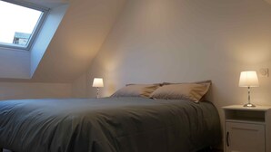 Un dormitorio con techo inclinado, una cama con colcha gris, dos lámparas de noche y un pequeño armario.