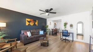 A modern living room with a dark wall, a ceiling fan, a brown leather sofa, a coffee table, and a dining area with chairs and a table.