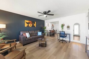 A modern living room with a dark wall, a ceiling fan, a brown leather sofa, a coffee table, and a dining area with chairs and a table.