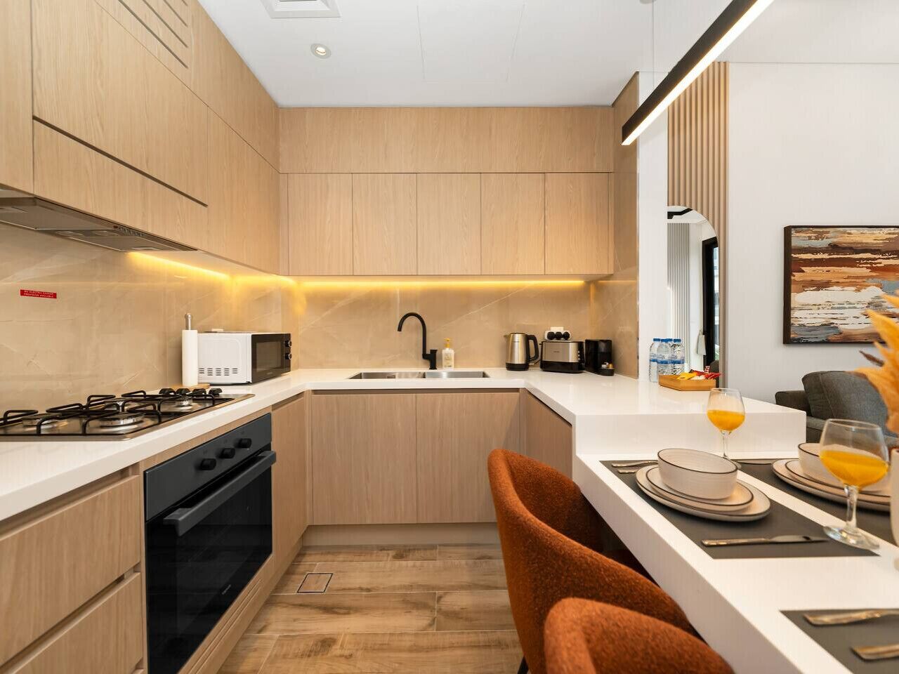 Cozinha moderna com armários de madeira, fogão a gás, micro-ondas e pia. Há uma área de refeições com uma mesa posta para dois e uma cadeira.