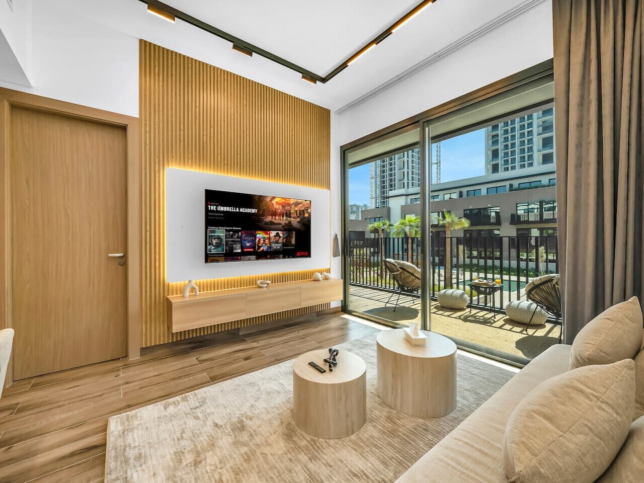 A modern living room with a large TV, wooden panel wall, and a balcony with outdoor furniture.