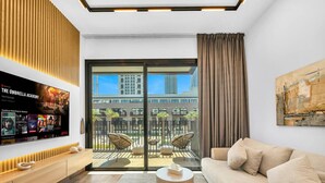 A modern living room with a sofa, coffee table, and TV. There's a balcony with chairs and a view of buildings. - Brand New Luxury 2Bedroom in JVC with 5* Amenities (Dubai)