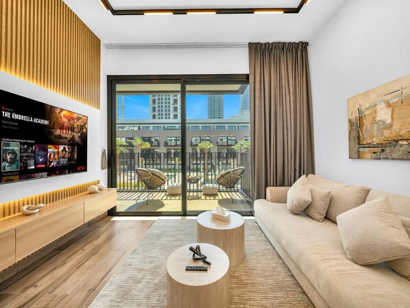 Ein modernes Wohnzimmer mit Sofa, Couchtisch und Fernseher. Es gibt einen Balkon mit Stühlen und Blick auf Gebäude.