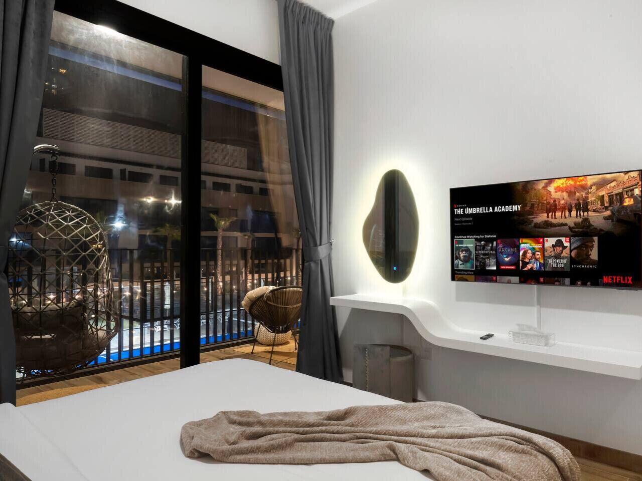 A modern hotel room with a large TV displaying The Umbrella Academy, a balcony with a pool, and a cozy seating area.