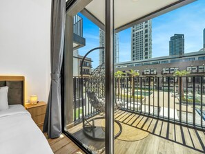 Ein Hotelzimmer mit Balkon, einem Bett, einem Nachttisch und Blick auf eine Stadtlandschaft sowie einen Poolbereich.