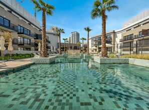 Ein modernes Hotel mit einem zentral gelegenen Schwimmbad, umgeben von Palmen und Liegestühlen.