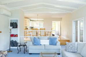 A spacious living area with a sofa set, a coffee table, and a floor lamp. A dining area with a table and chairs is visible in the background. - Soar Above the Sea at Eagle’s Perch Oceanfront Retreat (Nanoose Bay)