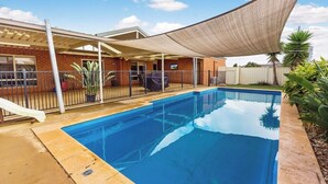 Una piscina con zona sombreada, un edificio de ladrillo y un patio cercado.